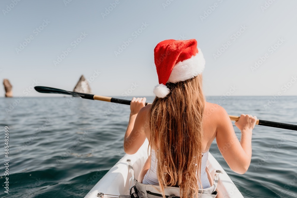 Obraz premium Woman in kayak back view. Happy young woman in Santa hat floating in kayak on calm sea. Summer holiday vacation and cheerful female people relaxing having fun on the boat.