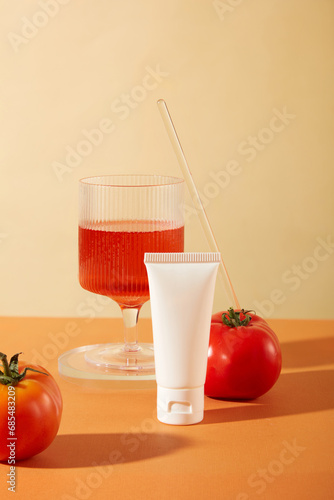 Front view of an unlabeled cosmetic tube and a glass of tomato juice on a beige and orange background. Tomatoes contain vitamin C which is an effective anti-oxidant.