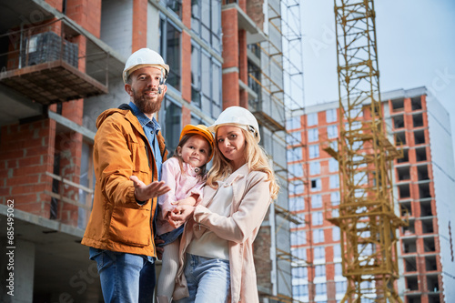 Man tossing up apartment keys and smiling while standing next to wife and daughter outside building under construction. Happy family homeowners posing on the street at construction site.