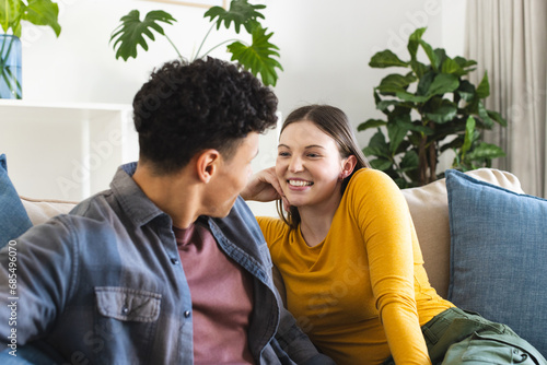 Happy diverse couple sitting on couch and looking at each other in living room at home, copy space