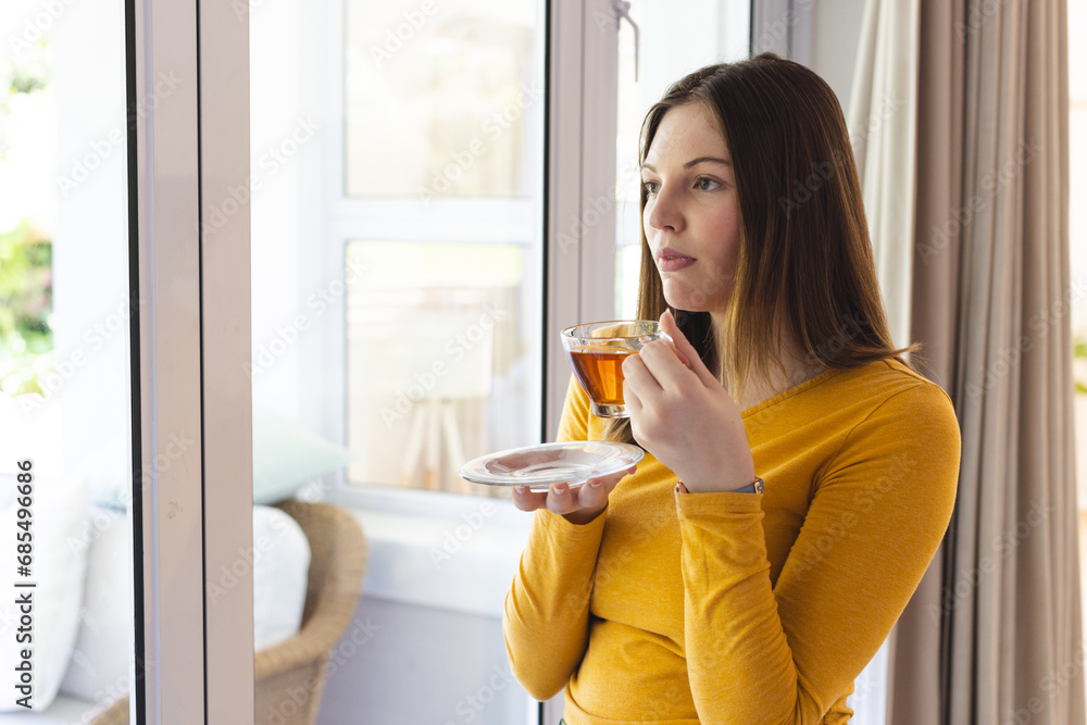 Thoughtful caucasian woman holding tea cup by window in living room at home, copy space Thoughtful caucasian woman holding tea cup by window in living room at home, copy space