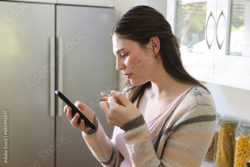 Focused caucasian woman standing  holding tea cup  using smartphone in kitchen at home  copy space