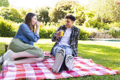 Happy diverse couple sitting on blanket, having picnic, drinking juice in sunny garden, copy space