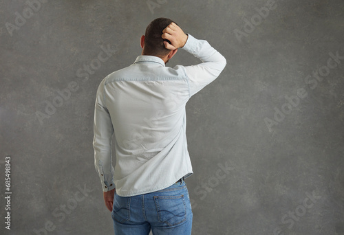 Unrecognizable man stands with his back to the camera and scratches the back of his head, not knowing what to do or what to choose. Confused man looks at the gray background. Place for text. photo