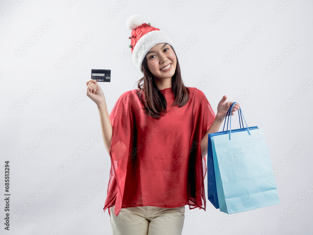Obraz premium A portrait of a happy Asian woman celebrating Christmas, wearing a red dress and Santa hat, carrying a shopping bag, and displaying a shopping card. Isolated against a white background.