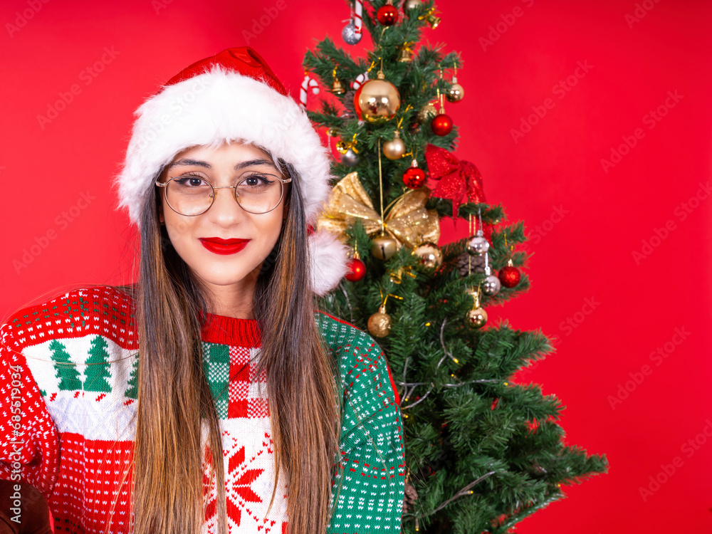 Santa woman, portrait of caucasian young attractive Santa woman. Wear hat, knitted sweater. Decorated Christmas tree, red studio background. Celebrating happy new year eve, noel concept idea.
