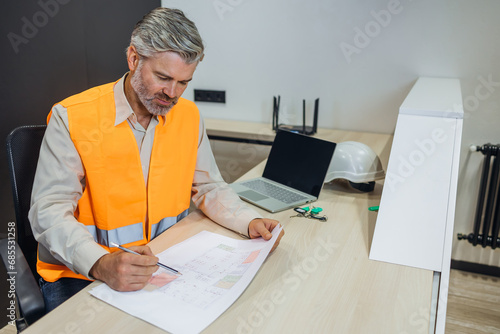 Man with Safety Orange Reflective Vest Sitting at Office Desk Working with Architect project. Senior Male Manager Doing Paperwork in engineer studio