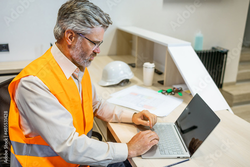 mature senior male sitting and using laptop at office factory. man worker in vest safety typing photo