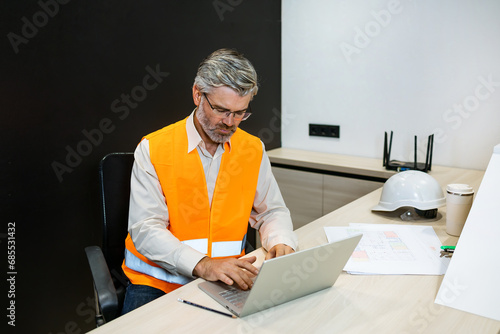 senior mature man engineer with laptop computer typing on laptop wear safety vest. Work housing projects and architecture