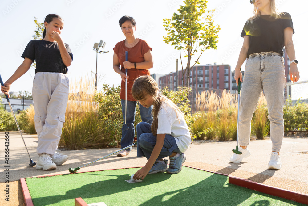 Cute school girl playing mini golf with family. Happy toddler child having fun with outdoor activity. Summer sport for children and adults, outdoors. Family vacations or resort. - obrazy, fototapety, plakaty 