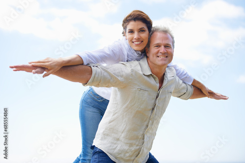 Mature couple, portrait and piggy back on vacation, airplane hands and happiness in blue skies. Man, woman and bonding together with smile, marriage and commitment with affection, holiday and joyful