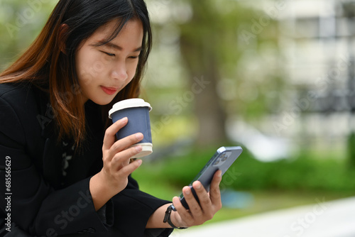 Happy young Asian woman drinking coffee and reading message on mobile phone