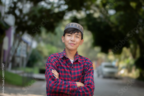 Portrait of young asian muslim or islamic schoolboy in red plaid shirt, standing and smilling in the middle way within his secondary school, soft and selective focus, happiness of humans concept.