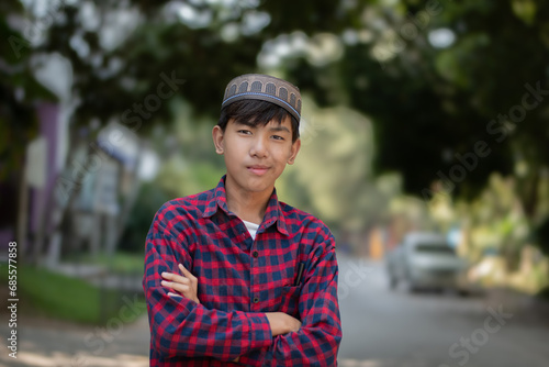 Portrait of young asian muslim or islamic schoolboy in red plaid shirt, standing and smilling in the middle way within his secondary school, soft and selective focus, happiness of humans concept.