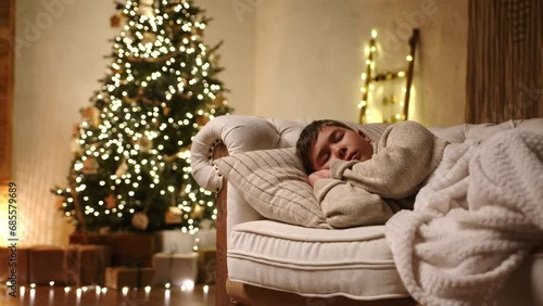 Young boy sleeping on modern light sofa, covered with plaid of milky color in a room with muffled light, plenty of gift-wrapped boxes under xmas tree. High quality 4k footage photo