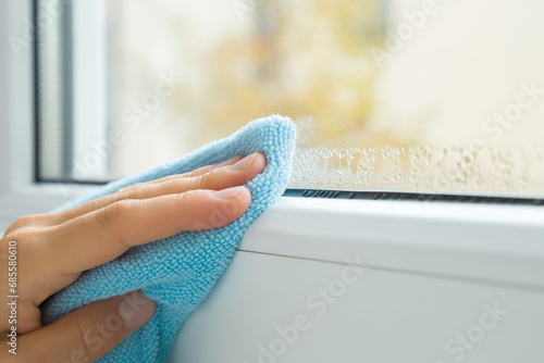 Young adult woman hand fingers holding dry blue soft microfiber rag and wiping water drops from glass and white window plastic frame. Closeup. Room humidity problem. photo
