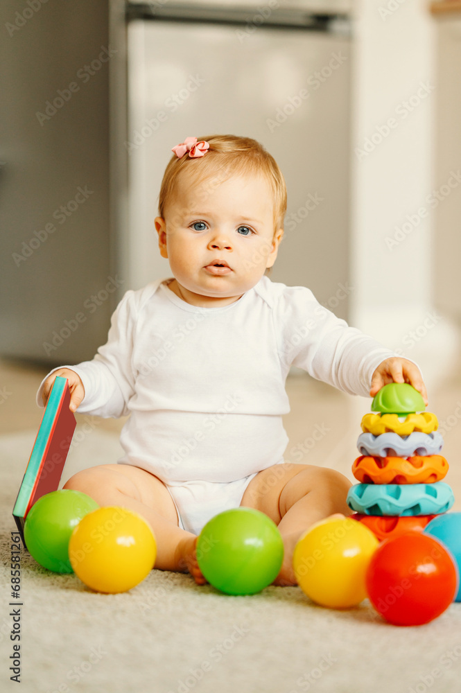 Obraz premium Vertical photo of baby girl sitting on floor with some toys.
