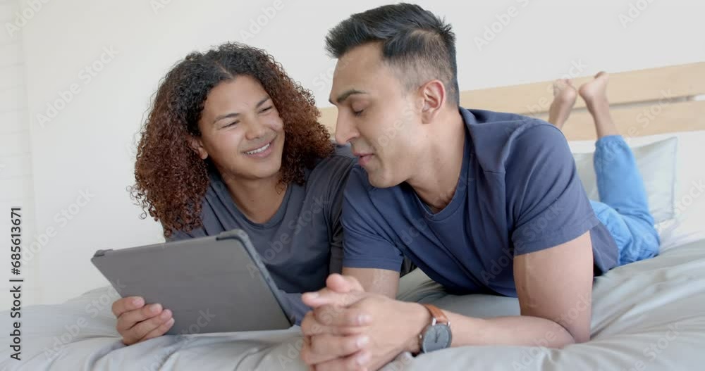 Happy diverse gay male couple lying on bed using tablet and talking in the morning, slow motion Happy diverse gay male couple lying on bed using tablet and talking in the morning, slow motion