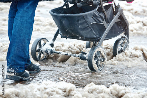 Woman push baby stroller through wet snow in winter season. Woman with baby carriage walk through snowy icy road after heavy snowfall in the city. Wheels of the baby stroller stuck on sleet photo