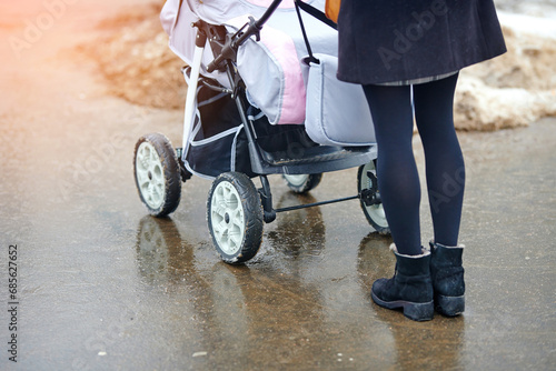Trendy woman with baby carriage walk on wet sidewalk in the city in winter season. Fashionable woman push baby stroller, walking on wet asphalt. Selective focus photo