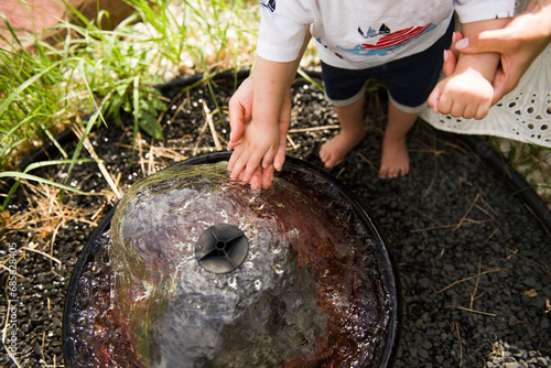 A mother's hands hold a boy's small hands in a decorative small circular garden fountain..