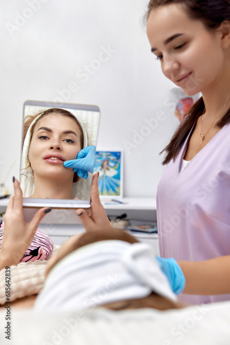 Beautiful woman looks at herself in the mirror after procedure lip augmentation in beauty medical clinic.