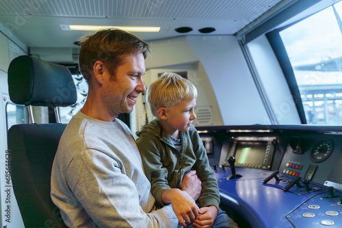 A man with European appearance of 40 years old and his blond son of 5 years old sit in the driver's cab of train