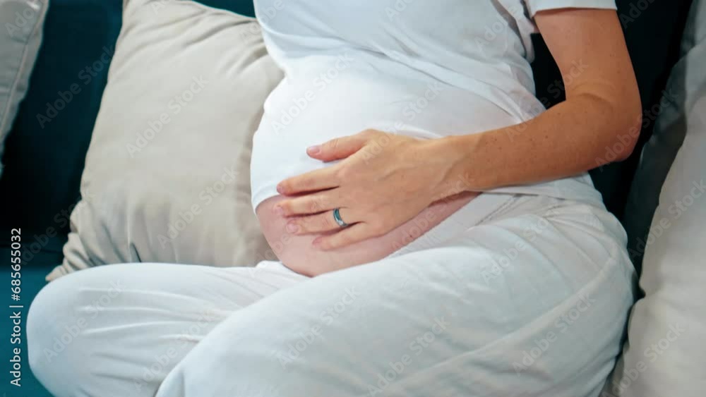 A young pregnant girl with a large stomach, sits at home on the sofa and touches her stomach with her hands. A young pregnant girl with a large stomach, sits at home on the sofa and touches her stomach with her hands.