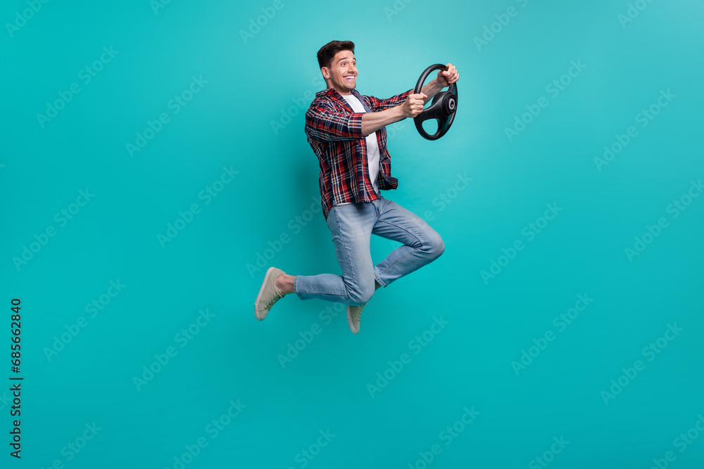 Full length photo of cheerful funky guy wear checkered shirt jumping high driving car isolated teal color background Full length photo of cheerful funky guy wear checkered shirt jumping high driving car isolated teal color background