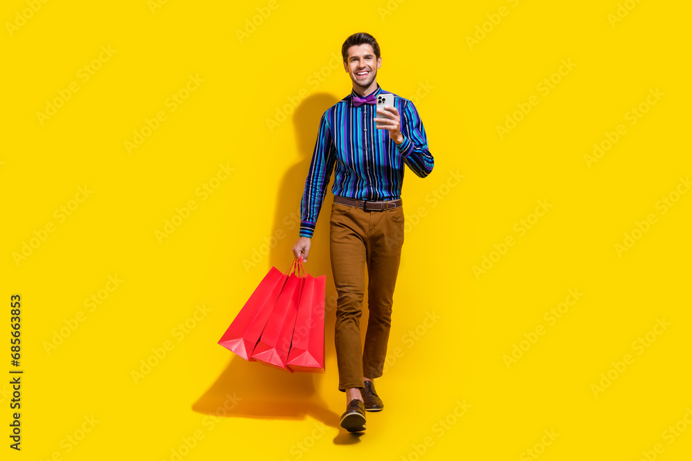 Full size photo of clever guy dressed striped shirt go with shopping bags order clothes on smartphone isolated on yellow color background Full size photo of clever guy dressed striped shirt go with shopping bags order clothes on smartphone isolated on yellow color background