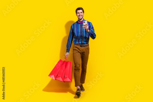 Full size photo of clever guy dressed striped shirt go with shopping bags order clothes on smartphone isolated on yellow color background