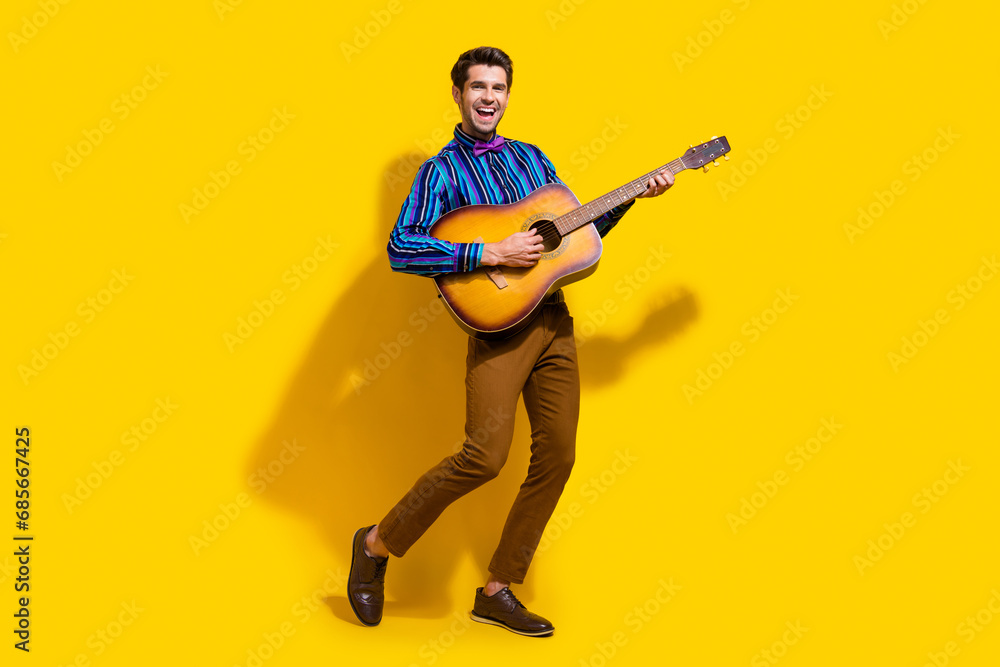 Full size photo of optimistic funky guy dressed striped shirt playing on acoustic guitar on stage isolated on yellow color background Full size photo of optimistic funky guy dressed striped shirt playing on acoustic guitar on stage isolated on yellow color background