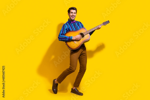 Full size photo of optimistic funky guy dressed striped shirt playing on acoustic guitar on stage isolated on yellow color background