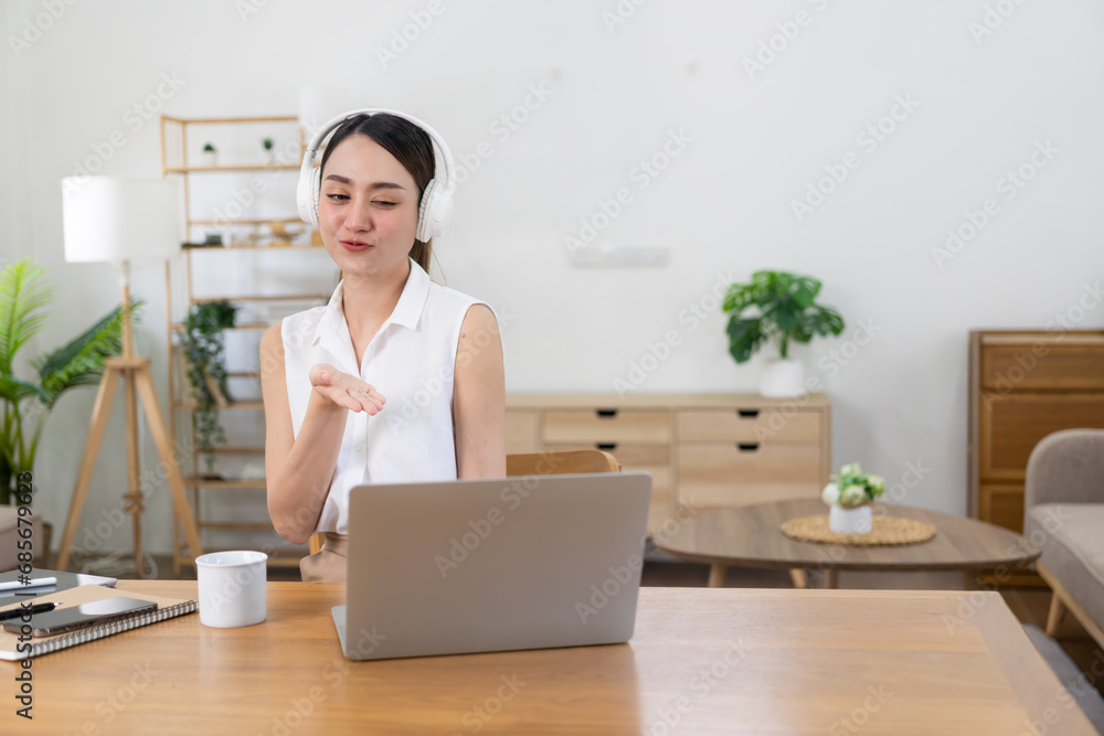 Smiling Asian girl wearing headphones using laptop Looking at the screen is happy listening to music while working online. Excited about student language learning, work at home concept Smiling Asian girl wearing headphones using laptop Looking at the screen is happy listening to music while working online. Excited about student language learning, work at home concept