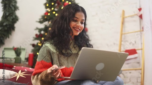 Pretty young indian woman sitting on bed paying with credit card on laptop near Christmas decoration at cozy home Happy customer enjoying online shopping on holidays sales indoors