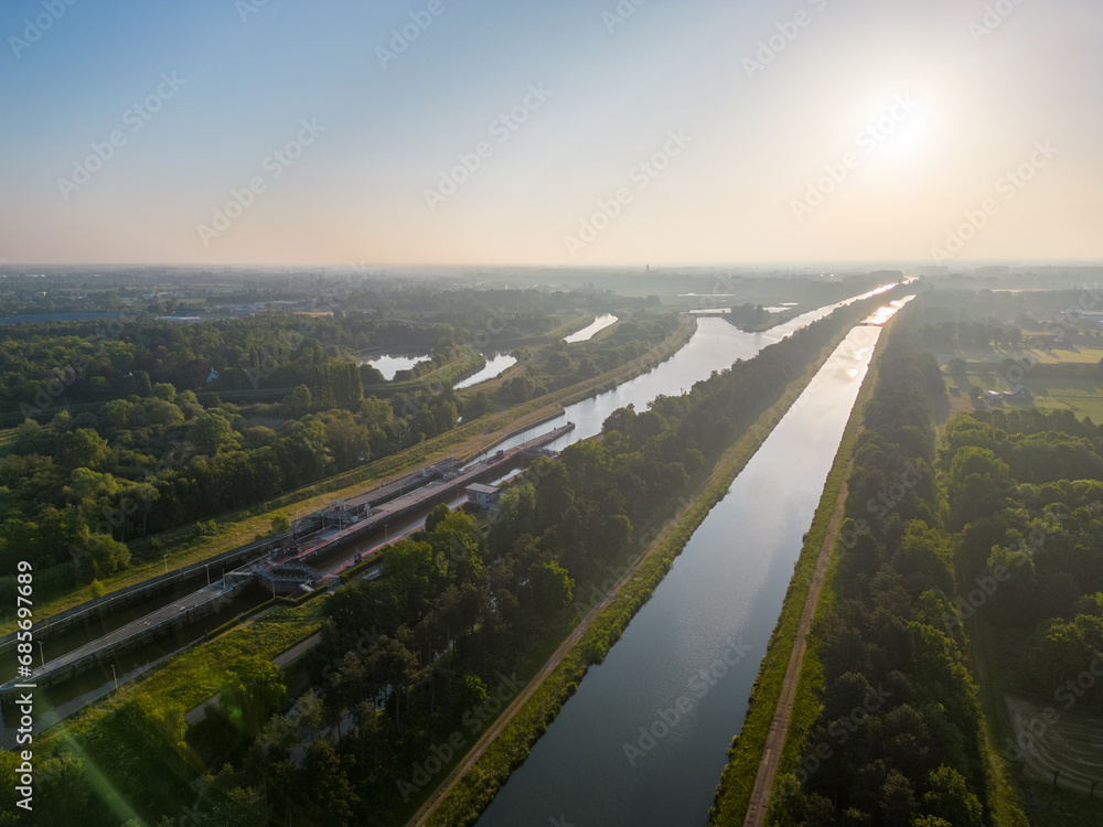 This image captures a breathtaking aerial view of a canal at sunrise, where the first golden rays of the day reflect off the still waters. Flanked by roads and lush greenery, the canal stretches into