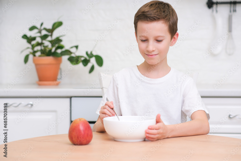 Little boy eating cereal with milk, apple for breakfast at wooden table in light white kitchen. Little boy eating cereal with milk, apple for breakfast at wooden table in light white kitchen.