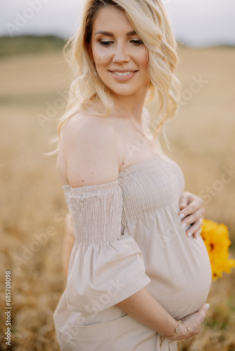 Portrait of a happy pregnant blonde woman with a straw basket and a bouquet of sunflowers spending time outdoors in a wheat field at sunset. The concept of motherhood.