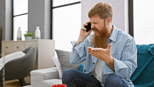 Unhappy young redhead man, argument escalating while using phone on sofa, expression shows disagreement, home setting amplifying problem, adds serious tone to indoor conversation photo