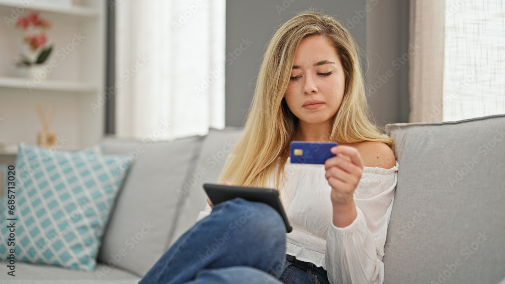 Young blonde woman shopping with touchpad and credit card sitting on sofa at home Young blonde woman shopping with touchpad and credit card sitting on sofa at home