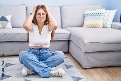Young caucasian woman sitting on the floor at the living room suffering from headache desperate and stressed because pain and migraine. hands on head.