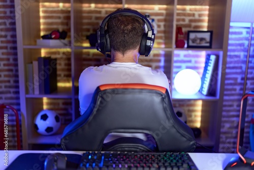 Middle age man with beard playing video games wearing headphones standing backwards looking away with crossed arms