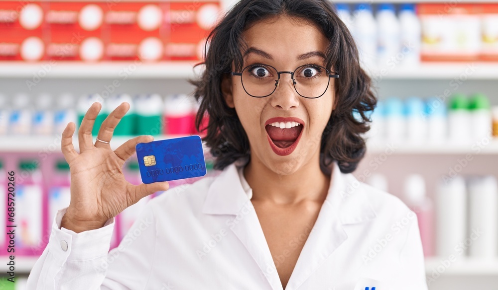 Young hispanic woman working at pharmacy drugstore holding credit card scared and amazed with open mouth for surprise, disbelief face