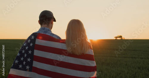 A couple of farmers with an American flag on their shoulders look at a wheat field where a tractor is working in the distance. photo