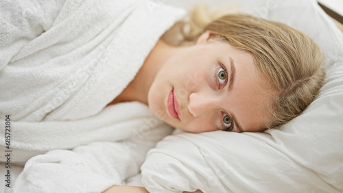 Young blonde woman wearing bathrobe hugging pillow with serious face at bedroom