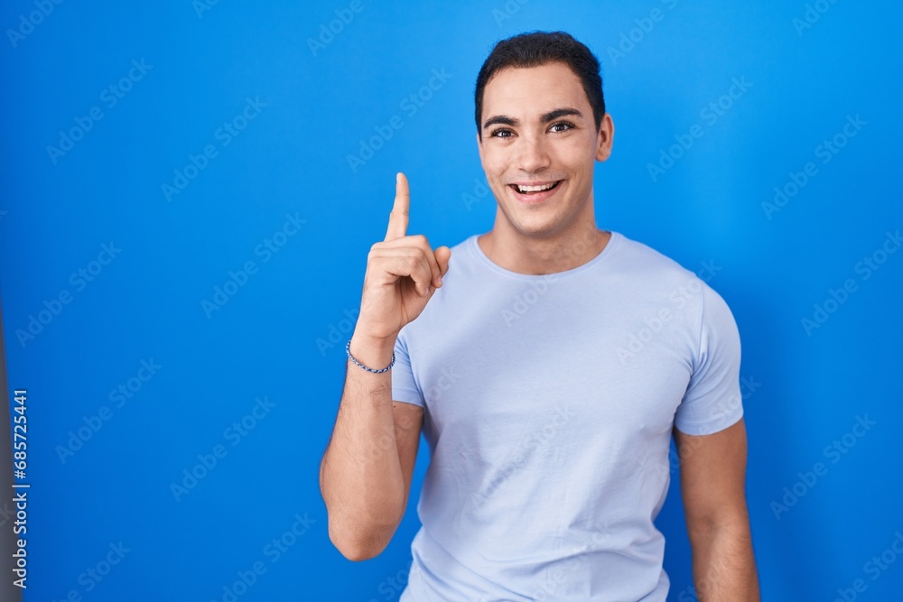 Young hispanic man standing over blue background pointing finger up with successful idea. exited and happy. number one. Young hispanic man standing over blue background pointing finger up with successful idea. exited and happy. number one.