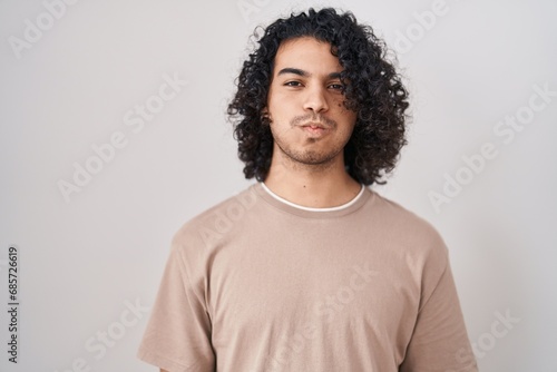 Hispanic man with curly hair standing over white background puffing cheeks with funny face. mouth inflated with air, crazy expression. photo