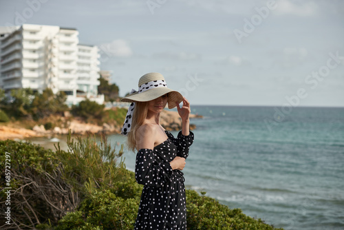 Beautiful young woman in hat and polka dot dress standing on seashore. Salou, Costa Dorada in province of Tarragona. Spain.