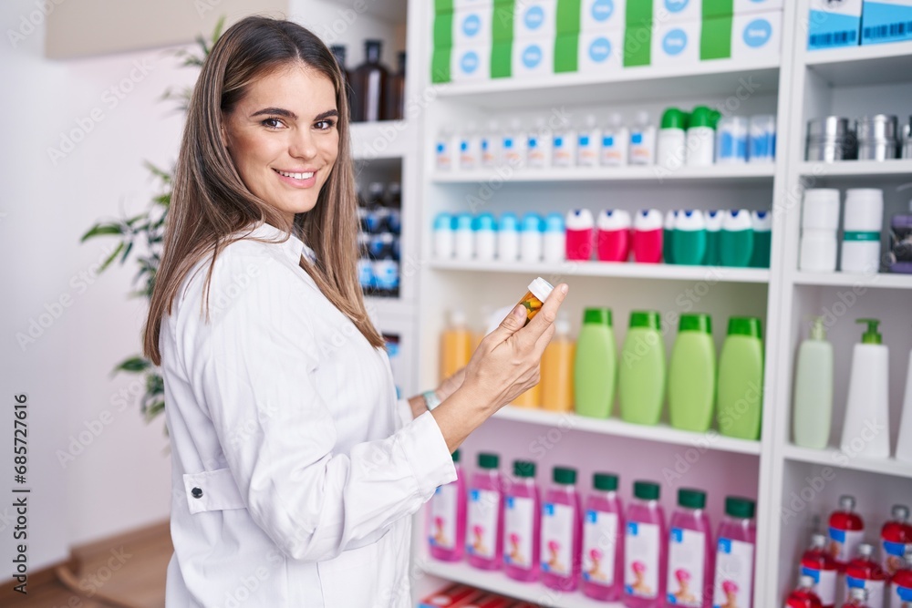 Young beautiful hispanic woman pharmacist smiling confident holding pills bottle of shelving at pharmacy