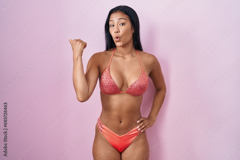Hispanic woman wearing bikini surprised pointing with hand finger to the side, open mouth amazed expression. Hispanic woman wearing bikini surprised pointing with hand finger to the side, open mouth amazed expression.